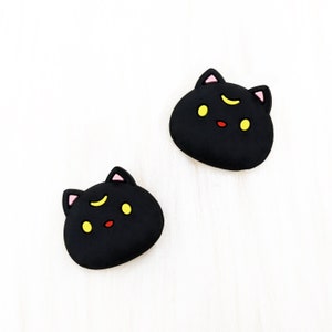 May include: Two black cat-shaped rubber charms with yellow eyes and pink ears. The cats have a crescent moon on their foreheads.