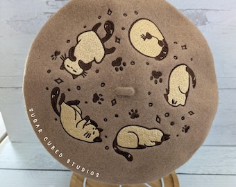 May include: A brown beret with a design of five cats in various poses, surrounded by paw prints and small diamonds. The text "SUGAR CUBED STUDIOS" is embroidered on the side.
