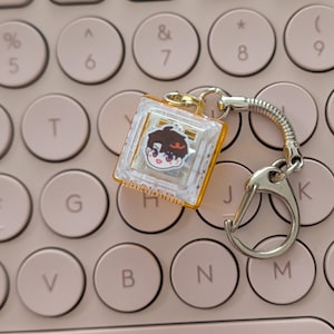 May include: A clear, square mechanical keyboard switch keychain with a gold-colored base. The switch features a small, illustrated cartoon face with brown hair and red eyes. The text SUGARCUBEDSTUDIOS is printed in small letters on the side.