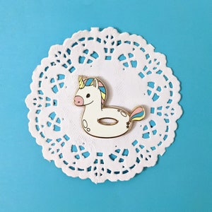 Può includere: Spilla in smalto dorato con un unicorno bianco con coda arcobaleno, all'interno di un anello gonfiabile bianco. La spilla è su un pizzo bianco.
