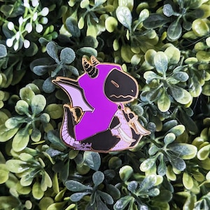 May include: Purple and black enamel pin of a dragon wearing a purple hooded cloak and holding a sword. The pin has a gold outline and the text "Sugar Cubed Studios" is printed on the bottom.