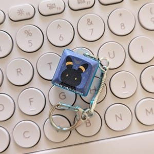 May include: A blue and black keycap charm with a cartoon character on it. The charm is attached to a silver keychain and is sitting on a white keyboard.