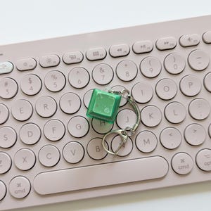 May include: A green square keyring with a silver key ring and chain rests on a light pink keyboard. The keyboard has round, white keys with black lettering. The keyring appears to be a miniature keyboard key.