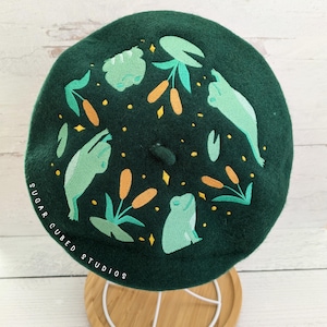 May include: A dark green beret with an embroidered design of frogs, lily pads, and cattails. The text "SUGAR CUBED STUDIOS" is embroidered on the side of the beret.