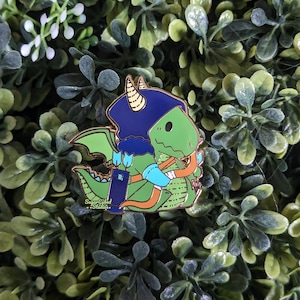 Puede incluir: Dragón verde con un sombrero azul y sosteniendo un arco y una flecha. El dragón es un pin de esmalte de estilo de dibujos animados.