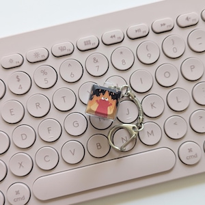 May include: A square keychain with a cartoon character design. The keychain features a red character with flames and the text "SUGAR CUBESTUDIOS". The keychain is attached to a silver keyring and is resting on a light pink keyboard.
