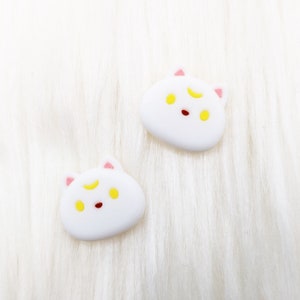 May include: Two white cat-shaped charms with pink ears and yellow eyes. Each charm has a crescent moon on its forehead.