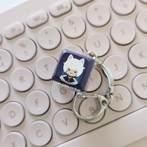 Op de afbeelding: Een paarse en witte keycap-sleutelhanger met een personage-illustratie en de tekst "SUGAR CUBED STUDIOS".