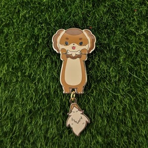 May include: A brown and cream enamel pin of a cartoon otter holding a fish in its paws. The otter has a brown body, cream coloured paws, and a brown head with cream coloured ears. The otter's tail is a separate charm that hangs from the pin.