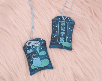 May include: Two blue fabric charms with a camouflage pattern and light blue cords. One charm features a frog on a lily pad, the other has Japanese text. The charms are designed with a night sky theme.