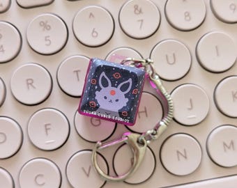 May include: A pink and purple key chain charm featuring a cartoon character with a white body, pink ears, and a red nose. The character is surrounded by a dark blue background with white stars and eyes. The charm is attached to a silver key chain with a silver ring. The text "SUGAR CUBED STUDIOS" is printed on the charm.