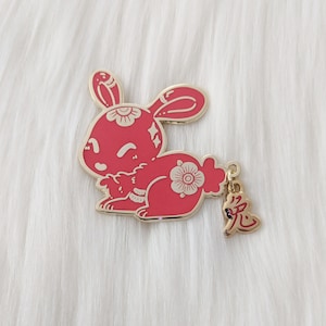May include: A red enamel pin featuring a stylized rabbit with white floral details. The rabbit is holding a smaller rabbit in its arms. The pin has a gold finish and a dangling charm with the Chinese character for "rabbit" in gold.