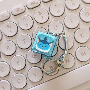 May include: A blue and white key chain with a cartoon character on a square charm. The charm has the text "SUGAR CUBED STUDIOS" on the bottom. The key chain is on a white keyboard.