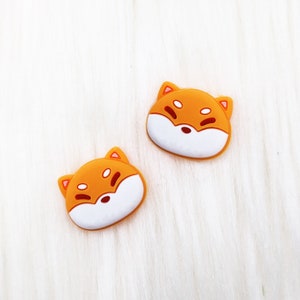 Op de afbeelding: Twee oranje en witte cartoon-honden gezichten met gesloten ogen. De honden gezichten zijn gemaakt van rubber en hebben een 3D-effect.