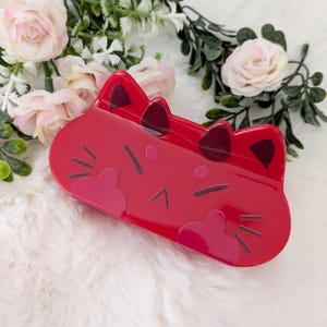 Puede incluir: Una pinza para el pelo roja con forma de gato y detalles negros. La pinza presenta un acabado brillante y un diseño lúdico, que incluye orejas de gato, bigotes y una nariz. Un accesorio divertido para cualquier peinado.
