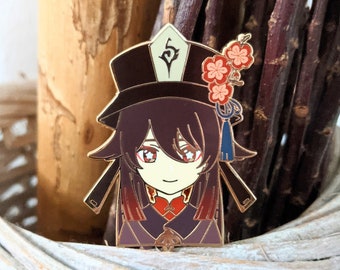 Op de afbeelding: Een gouden emaille pin met een cartoonfiguur met lang donker haar, dat een zwarte hoed draagt met een rood bloemenontwerp. Het personage draagt een rood-paarse outfit en heeft een licht gezicht met rode ogen.
