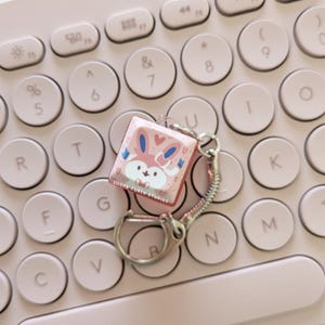 May include: A pink square keychain with a cartoon bunny design, hearts, and the text "SUGAR CUBED STUDIOS." The keychain is attached to a silver keyring and a pink chain, resting on a light pink keyboard.