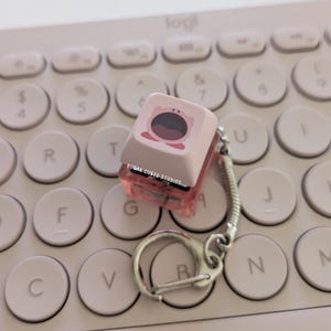Puede incluir: Un llavero rosa y transparente con un personaje rosa de boca grande y el texto "SUGAR CUBED STUDIOS". El llavero está sobre un teclado blanco.