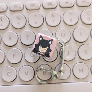 Könnte beinhalten: Ein rosa-weißer Keycap mit einer Comicfigur, die ein schwarz-weißes Outfit und rosa Ohren trägt. Der Keycap ist an einem silbernen Schlüsselanhänger mit einem federbelasteten Verschluss befestigt. Der Keycap befindet sich auf einer weißen Tastatur mit runden Tasten.