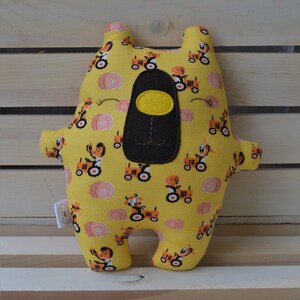 May include: A yellow plush bear-shaped pillow with a repeating pattern of orange tractors and red and white swirls. The bear has a black nose and a yellow oval accent. The pillow has small arms and legs.