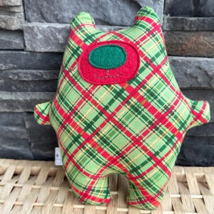 May include: A handmade plush toy in a whimsical design. The toy features a green and red plaid pattern with a red and green felt face. It has small arms and legs, and two small ears. The toy is sitting on a woven surface.