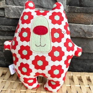 May include: A handmade plush bear with a red and white floral pattern. The bear has a light green face, a red nose, and embroidered details. The bear is approximately 10 inches tall and has small arms and legs.