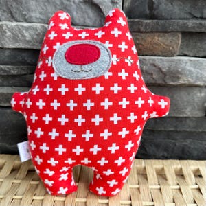 May include: A stuffed plush toy in the shape of a bear, featuring a red body with white cross patterns. The bear has a gray face with a red nose and small ears. The toy has small arms and legs.