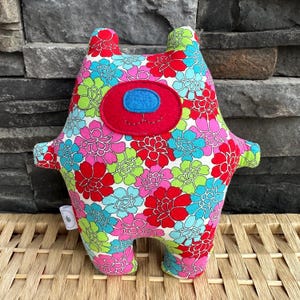 May include: A colorful plush toy in the shape of a creature with a floral pattern in red, pink, blue, and green. It has a red face with a blue oval and stitched details. The toy has small arms and legs.