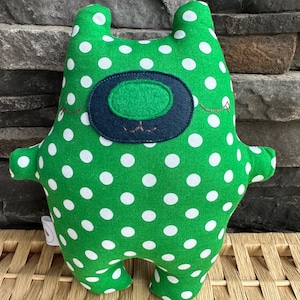 May include: A green plush toy in the shape of a creature with white polka dots. The toy has a dark blue and green oval-shaped face with embroidered details. It has small arms and ears.