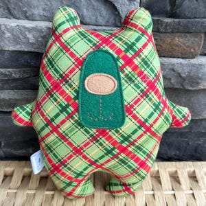 May include: A handmade plush toy in the shape of a bear, featuring a green, red, and cream plaid pattern. The bear has a green felt face with a tan oval nose and stitched details. The toy has small arms and legs.
