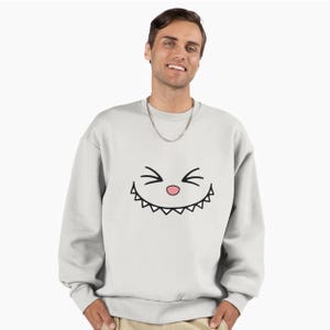 Puede incluir: Sudadera gris claro con un dise&ntilde;o de cara de dibujos animados. El dise&ntilde;o incluye l&iacute;neas negras para los ojos y la boca, y un peque&ntilde;o c&iacute;rculo rosa para la nariz. La sudadera es de cuello redondo.