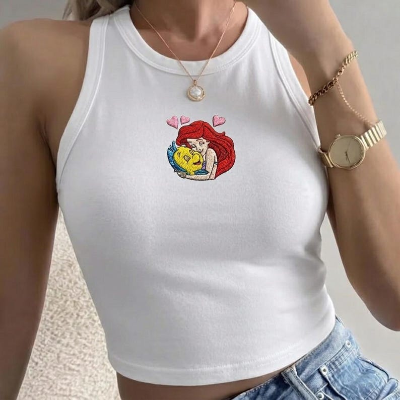 May include: White vest top featuring an embroidered image of Ariel from The Little Mermaid embracing Flounder, with two pink hearts. The top has a round neck and is sleeveless.