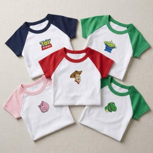 May include: Five short-sleeve raglan t-shirts with white bodies and colored sleeves. One shirt features the Toy Story logo, while the others display Woody, a green alien, a pink pig, and a green dinosaur character graphic on the front.