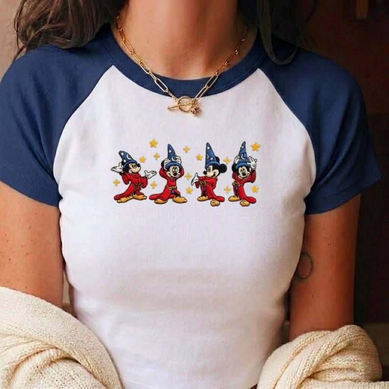 May include: A white raglan t-shirt with dark blue sleeves featuring a graphic print of four Mickey Mouse characters dressed as sorcerers in red robes and blue hats, surrounded by small yellow stars, paired with a gold-toned chain link necklace.