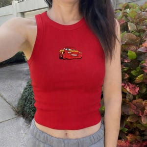 May include: A red ribbed tank top featuring a cartoon image of a red and yellow race car. The sleeveless top has a round neckline and is made of a soft, stretchy material. The car graphic is embroidered on the front.