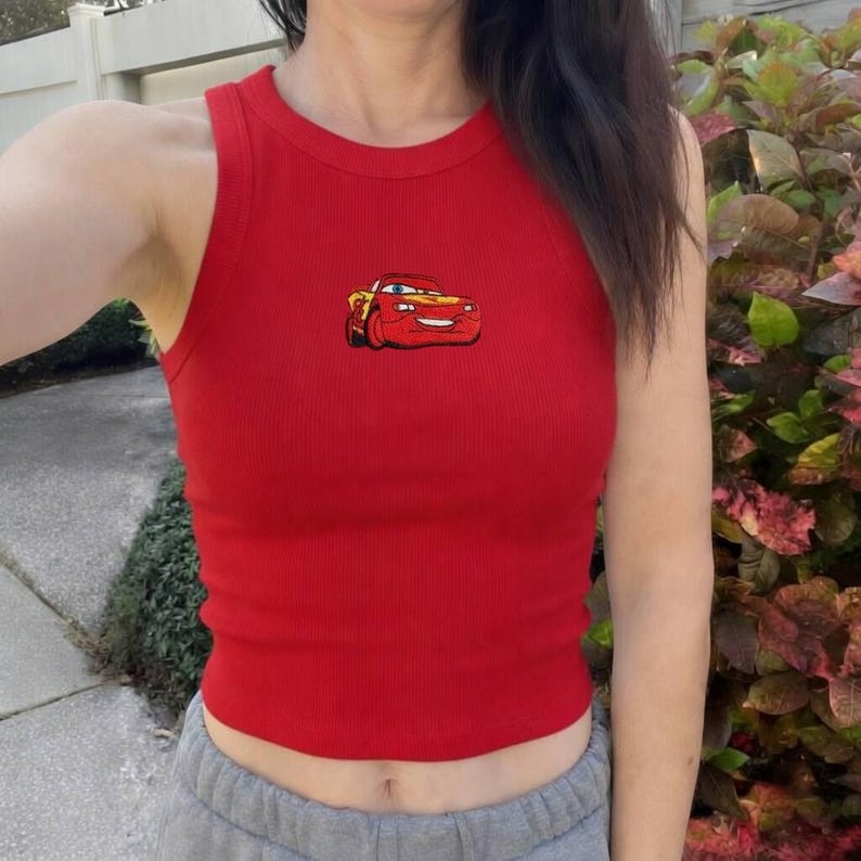 May include: A red ribbed tank top featuring a cartoon image of a red and yellow race car. The sleeveless top has a round neckline and is made of a soft, stretchy material. The car graphic is embroidered on the front.