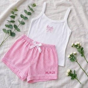 Puede incluir: Conjunto de pijama de camiseta sin mangas blanca y pantalones cortos a rayas rosas. La camiseta sin mangas tiene la palabra "bride" bordada en rosa con un lazo. Los pantalones cortos tienen un lazo blanco y la fecha "10.19.25" bordada en rosa. El conjunto se muestra sobre una superficie blanca.