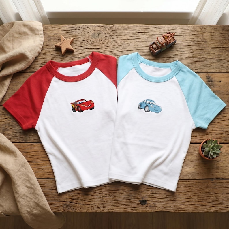 May include: Two short-sleeve raglan t-shirts with white bodies. One shirt features red sleeves and a red collar with a red race car graphic. The other shirt features light blue sleeves and a light blue collar with a light blue car graphic.