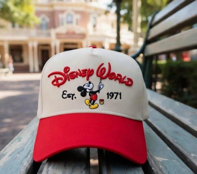 Op de afbeelding: Een baseballcap met een rode klep en een cr&egrave;mekleurige kroon. De cap heeft de woorden "Disney World" in het rood, samen met een afbeelding van Mickey Mouse en het jaar "1971".