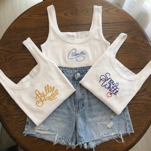 May include: Three white vest tops with embroidered text and a pair of denim shorts. One top says "Cinderella" in blue script, another says "Belly" in gold script, and the third says "Snow White" in blue script with a red apple.