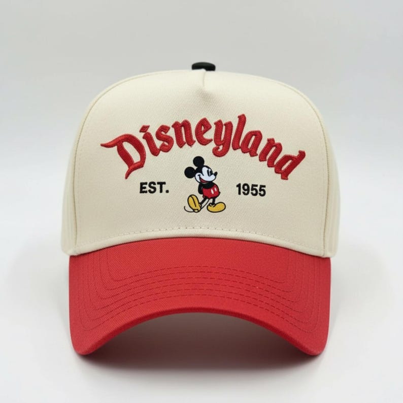 May include: A cream-colored baseball cap with a red brim. The cap features the word "Disneyland" in red script, along with a Mickey Mouse graphic and the text "EST. 1955".