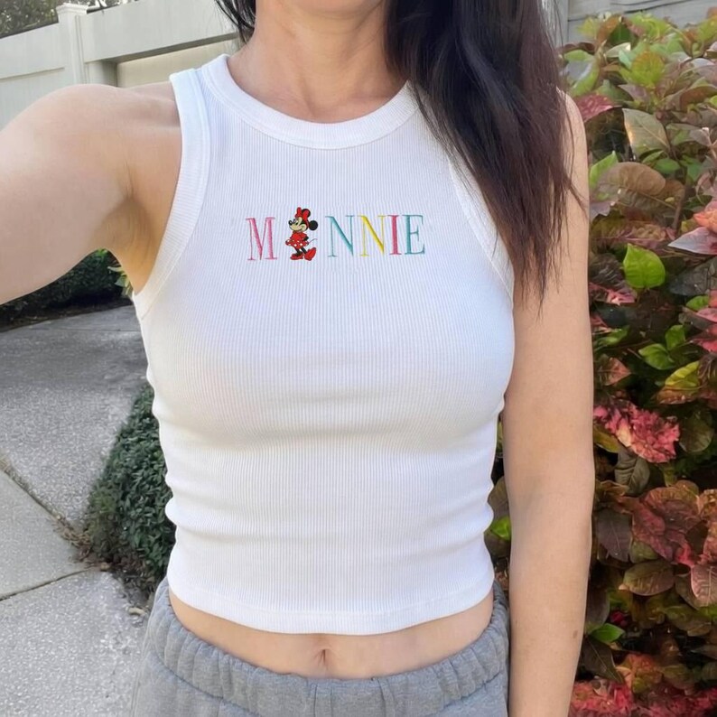 Puede incluir: Camiseta sin mangas blanca acanalada con la palabra "Minnie" bordada en letras coloridas. La "i" de Minnie se reemplaza con una imagen de dibujos animados de Minnie Mouse. La camiseta sin mangas es sin mangas y tiene un cuello redondo.