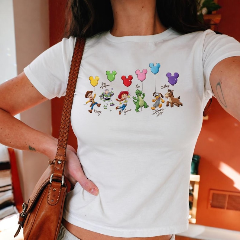 May include: White t-shirt featuring a colorful graphic of Toy Story characters holding Mickey Mouse-shaped balloons. The characters include Woody, Buzz Lightyear, Jessie, Rex, Slinky Dog, and Bullseye. The shirt is a crew neck style.