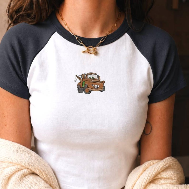 May include: A white raglan t-shirt with dark grey sleeves and a crew neckline. The centre of the chest features a small, detailed embroidery of a brown tow truck character with green accents, large eyes, and a wide, toothy smile.