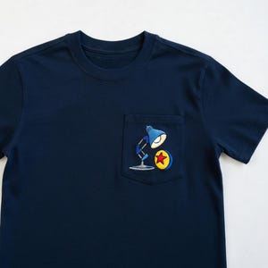 Op de afbeelding: Marineblauw T-shirt met een zakje met een geborduurd ontwerp van een blauwe bureaulamp, een gele cirkel en een rode ster. Het shirt heeft een ronde hals en korte mouwen.