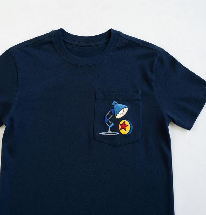Op de afbeelding: Marineblauw T-shirt met een zakje met een geborduurd ontwerp van een blauwe bureaulamp, een gele cirkel en een rode ster. Het shirt heeft een ronde hals en korte mouwen.