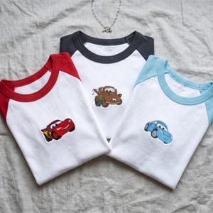 May include: Three raglan t-shirts with white bodies and coloured sleeves. The left shirt has red sleeves and an embroidered red race car. The centre shirt has dark grey sleeves and an embroidered brown tow truck. The right shirt has light blue sleeves and an embroidered light blue car.