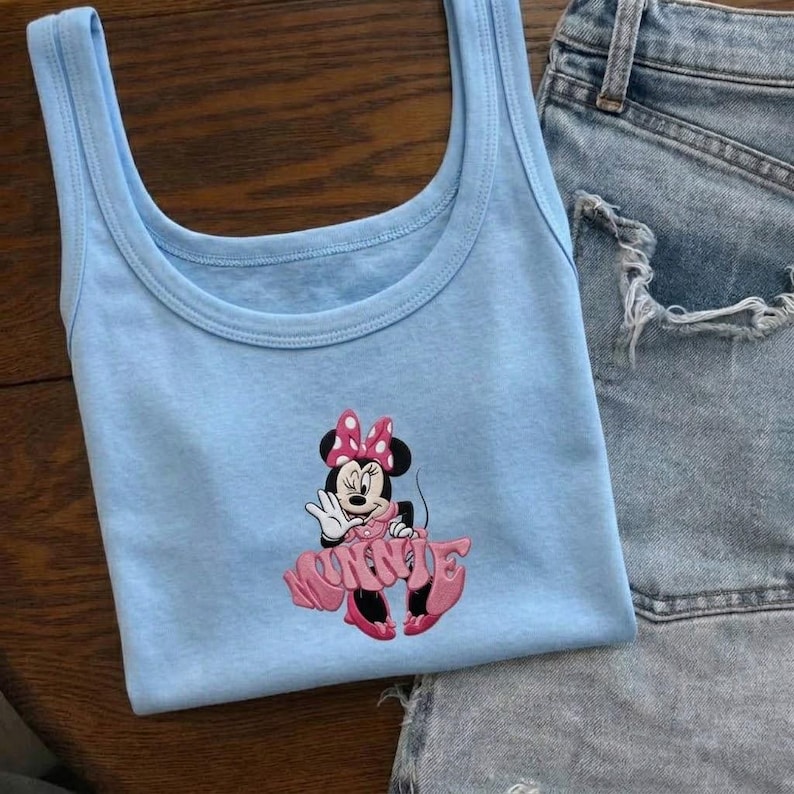Może przedstawiać: Jasnoniebieski top bez rękaw&oacute;w z haftowanym wzorem Myszki Minnie. Wz&oacute;r zawiera twarz Myszki Minnie, r&oacute;żową kokardę i napis "Minnie" w r&oacute;żowym skrypcie. Top bez rękaw&oacute;w jest zestawiony z dżinsowymi szortami.
