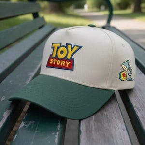 Op de afbeelding: Een baseballpet met een crèmekleurige kroon en een groene klep. De voorkant heeft het "Toy Story" logo in gele, blauwe en rode borduursels. Een kleine cartoon lamp en bal afbeelding staat aan de zijkant.