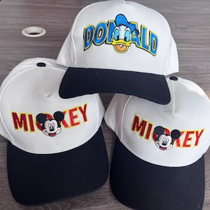 May include: Three white baseball caps with black brims. One cap features "DONALD" in blue lettering with Donald Duck's face. The other two caps display "MICKEY" in red lettering with Mickey Mouse's face. All caps have a classic, structured design.